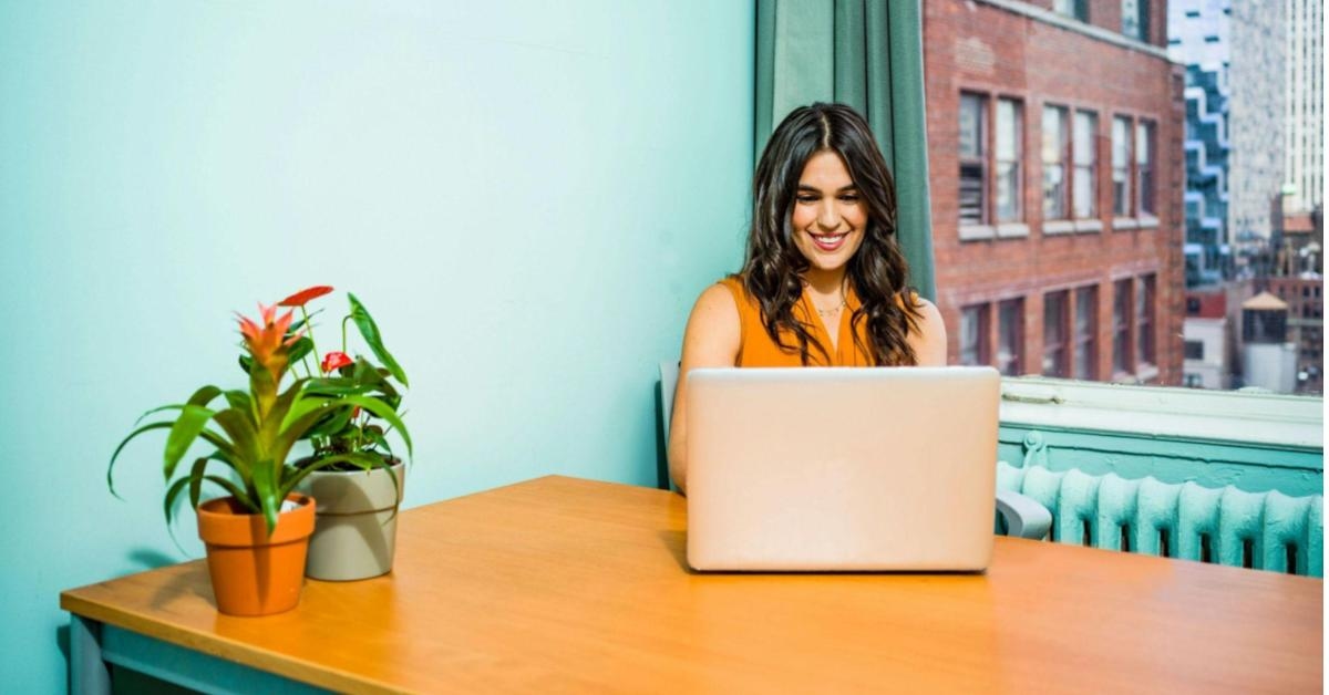 imagem mostra uma mulher, de cabelos longos e pretos, vestindo uma blusa laranja, sentada de frente para seu notebook cinza. Na imagem, é como se estivéssemos de frente para ela, que sorri enquanto observa algo na tela, uma alusão ao momento em que empreendedores buscam informações para se prevenir de ataques cibernéticos. A sala em que a mulher está tem paredes azuis claras, e podemos ver, por meio de uma janela, um prédio de tijolos.