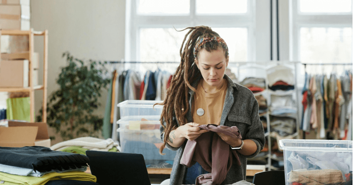 A imagem mostra uma mulher num estoque de loja de roupas. Ela é branca, usa camisa xadrez vermelha e está conferindo um documento. Uma alusão ao momento que empreendedores fazem balanços para compreender a saúde financeira do negócio.